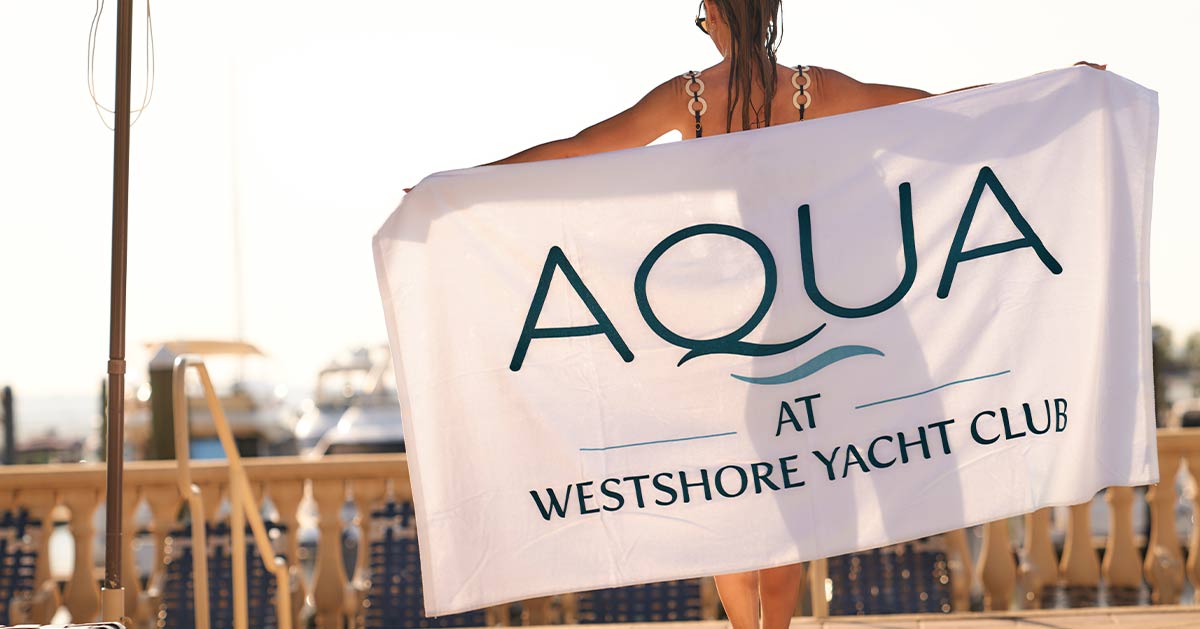 Woman holding a white beach towel with the AQUA logo on The Bay Club pool deck, with the marina in the background.